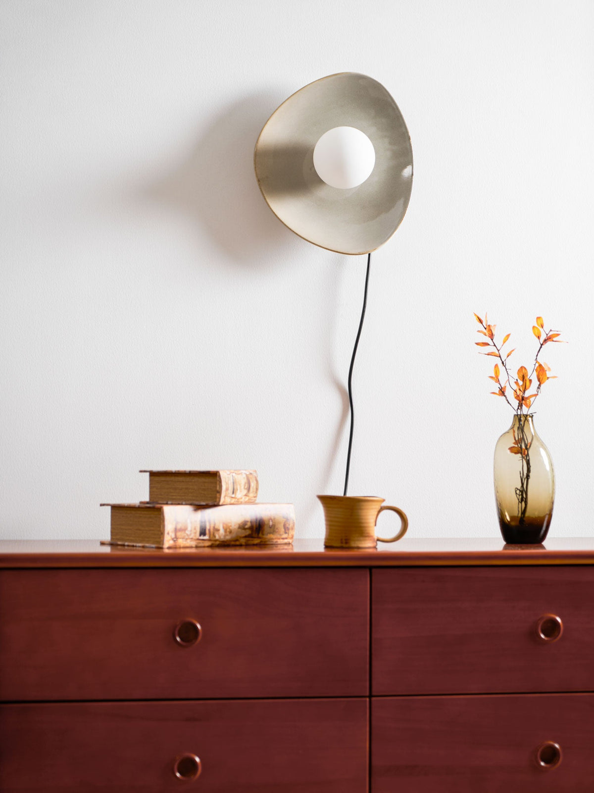 Unlit, a taupe Clérachic ceramic wall light with a white globe hangs above a dark wood chest of drawers, a cup and a vase of orange leaves.