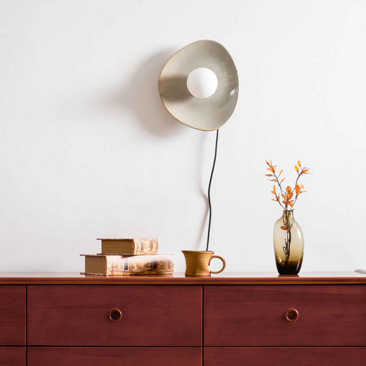 A taupe Clérachic ceramic wall light and white globe, switched off, above a wooden chest with stacked books and a glass vase.