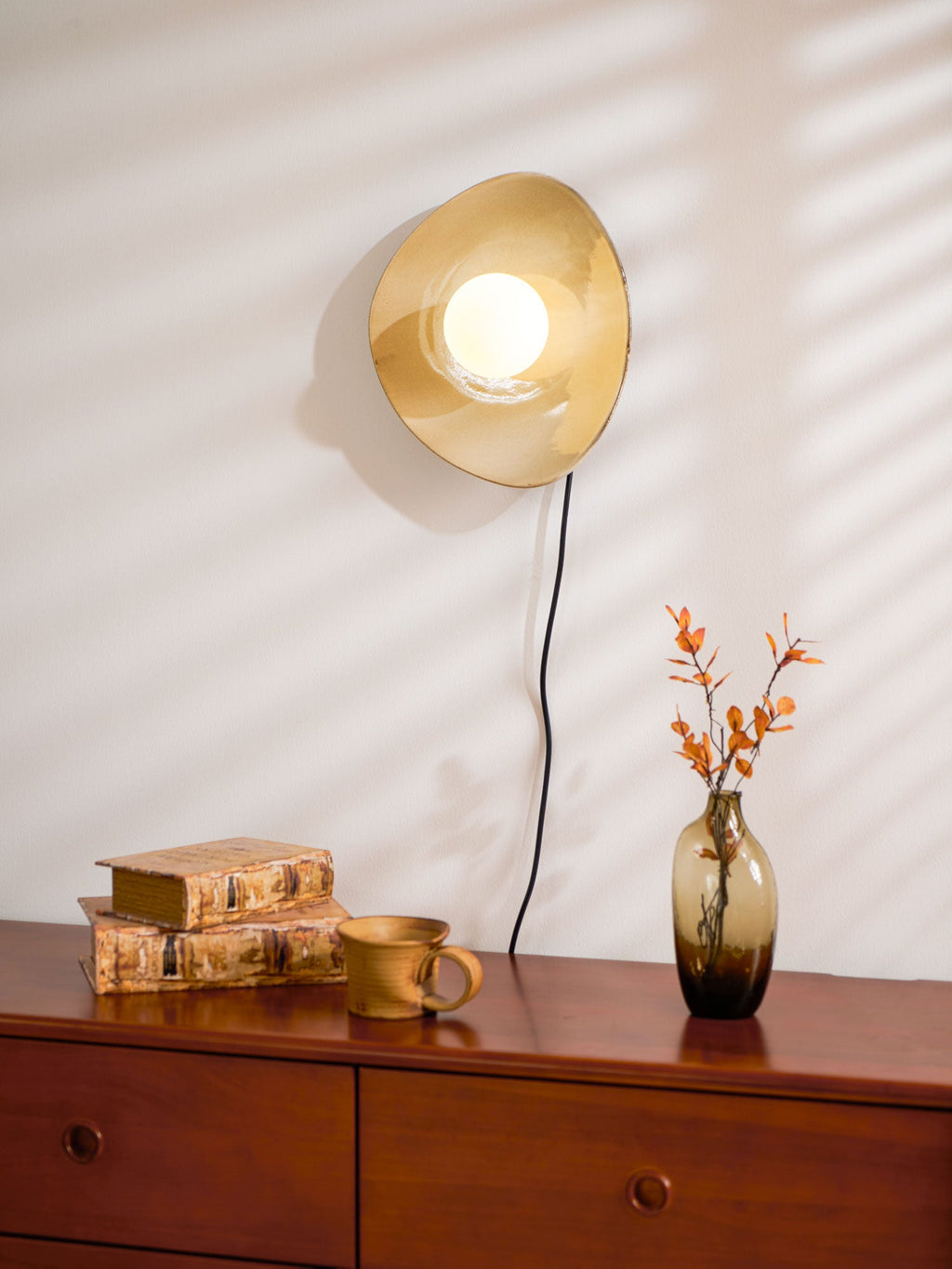 Stripes of sunlight fall across the white wall beside this lit Clérachic ceramic wall light, above a chest, books and a vase.
