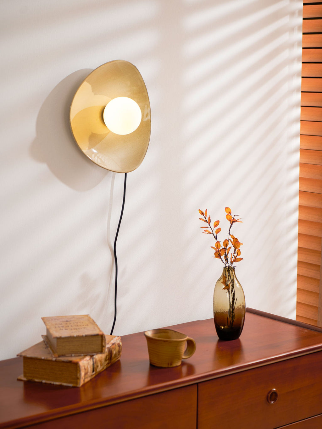 Window blinds throw light across the wall behind this lit Clérachic ceramic wall light, with a vase and books on the chest below.