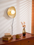 Window blinds throw light across the wall behind this lit Clérachic ceramic wall light, with a vase and books on the chest below.