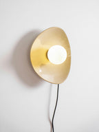 Close view of the lit globe and the golden glaze inside the ceramic disc on this Clérachic wall light, with a black cord below.