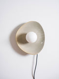 Front view of a taupe Clérachic ceramic wall light with a white globe bulb, its glazed edge catching the light and a black cord below.
