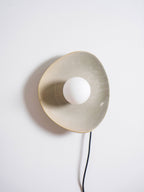 Front view of a taupe Clérachic ceramic wall light with a white globe bulb, its glazed edge catching the light and a black cord below.