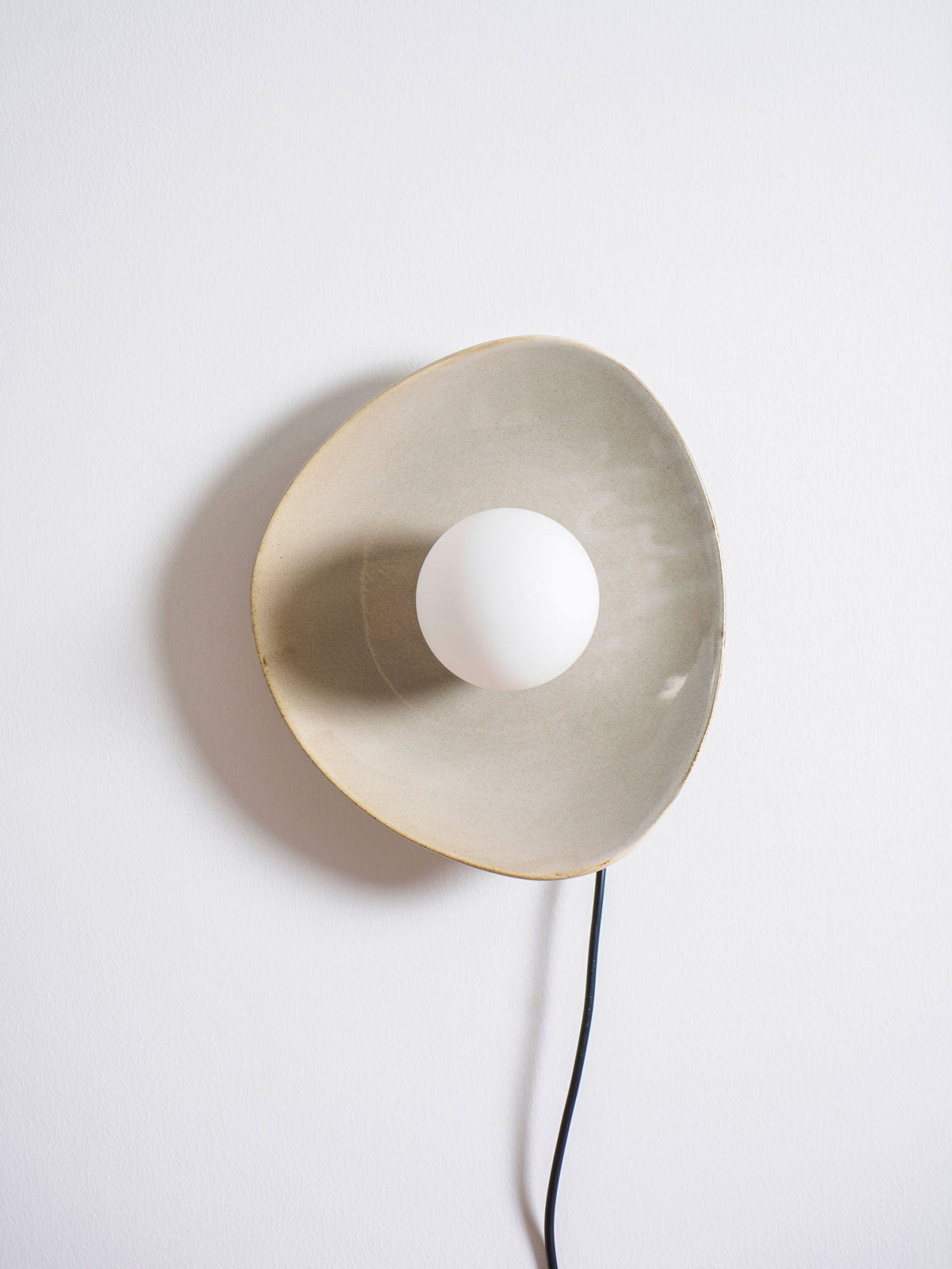 Front view of a taupe Clérachic ceramic wall light with a white globe bulb, its glazed edge catching the light and a black cord below.