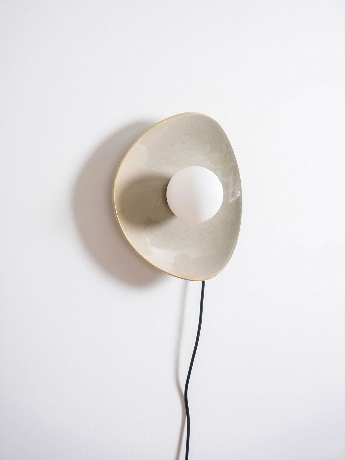 Angled view of this Clérachic taupe ceramic wall light and white globe, with a soft shadow on the wall behind and a black cord trailing down.
