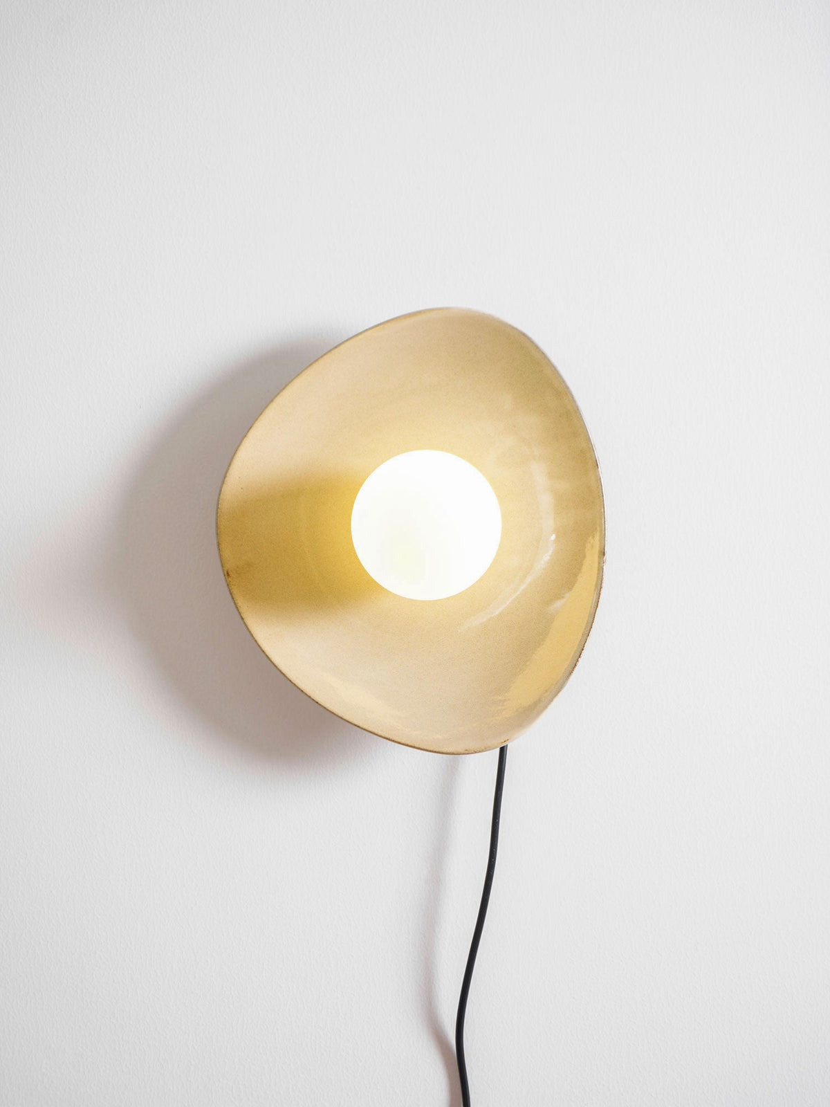 Lit, this Clérachic ceramic wall light shows a warm golden glow inside its disc and a soft shadow on the white wall.