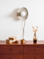 A taupe Clérachic ceramic wall light and its black cord hang above a wooden chest of drawers, with books, a mug and a vase of orange leaves.