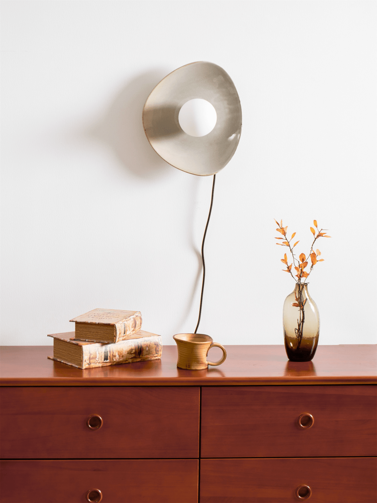 A taupe Clérachic ceramic wall light and its black cord hang above a wooden chest of drawers, with books, a mug and a vase of orange leaves.