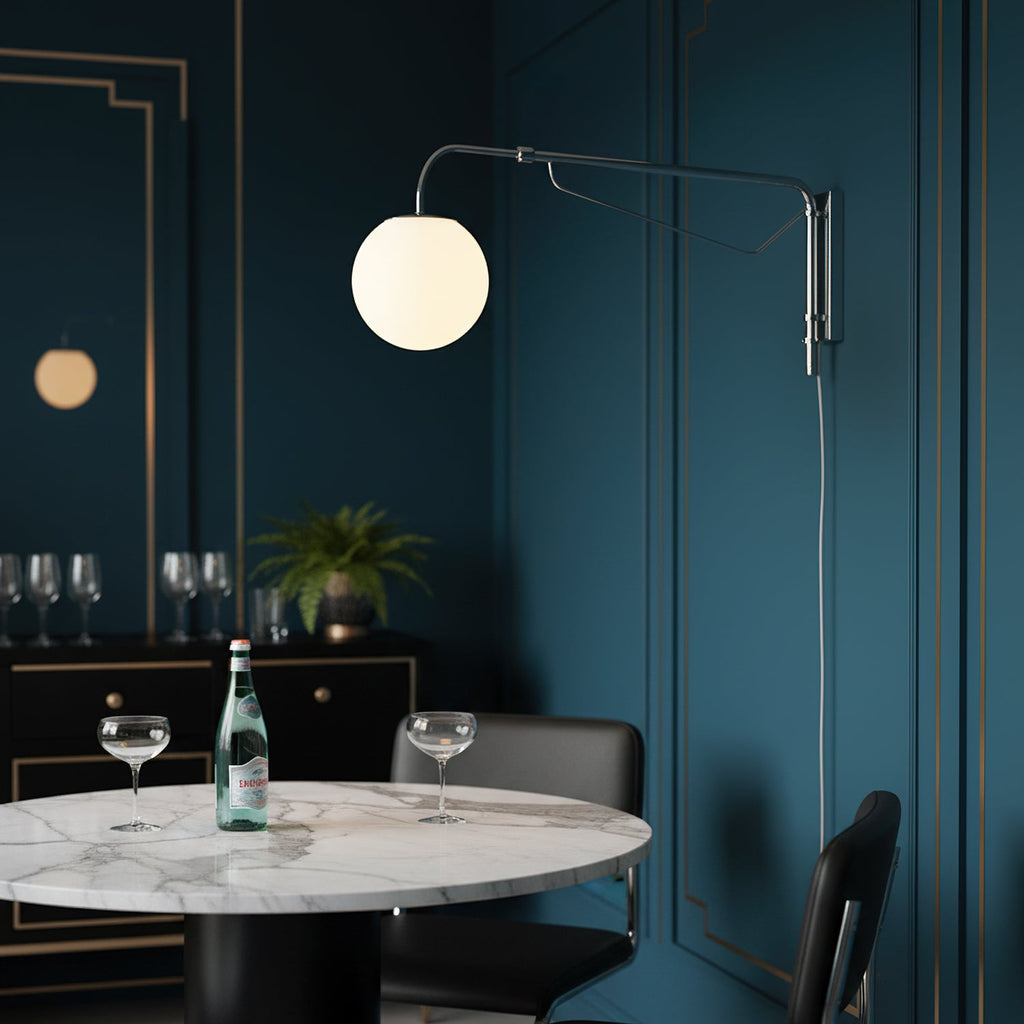 This Clérachic wall light with a white globe and chrome arm on a deep teal panelled wall, above a marble table with coupe glasses.