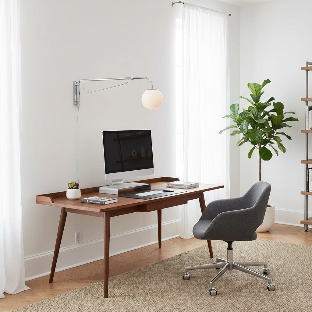A Clérachic plug-in wall light with a chrome arm hangs above a walnut desk with a monitor, a grey chair and a tall plant.