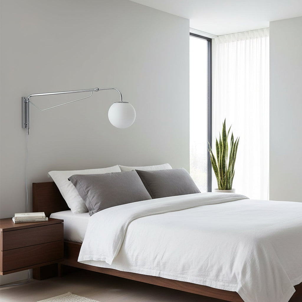 A chrome Clérachic wall light with a white globe above a bed with white linen, grey cushions and a walnut bedside table.