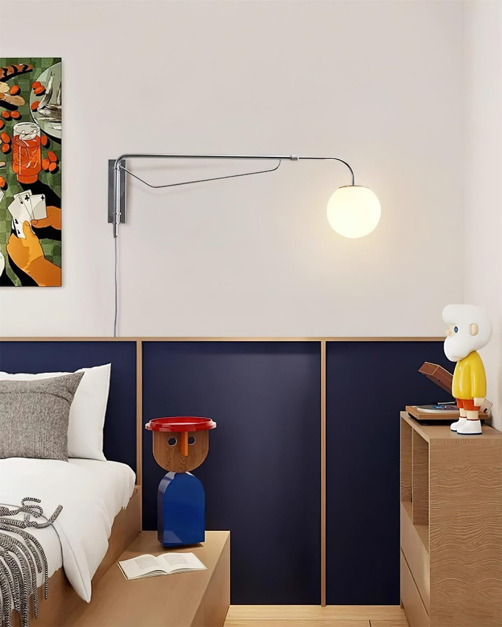 Chrome Clérachic wall light with a white globe lit on a pale wall above a navy panelled headboard and colourful figurines.