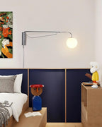 Chrome Clérachic wall light with a white globe lit on a pale wall above a navy panelled headboard and colourful figurines.