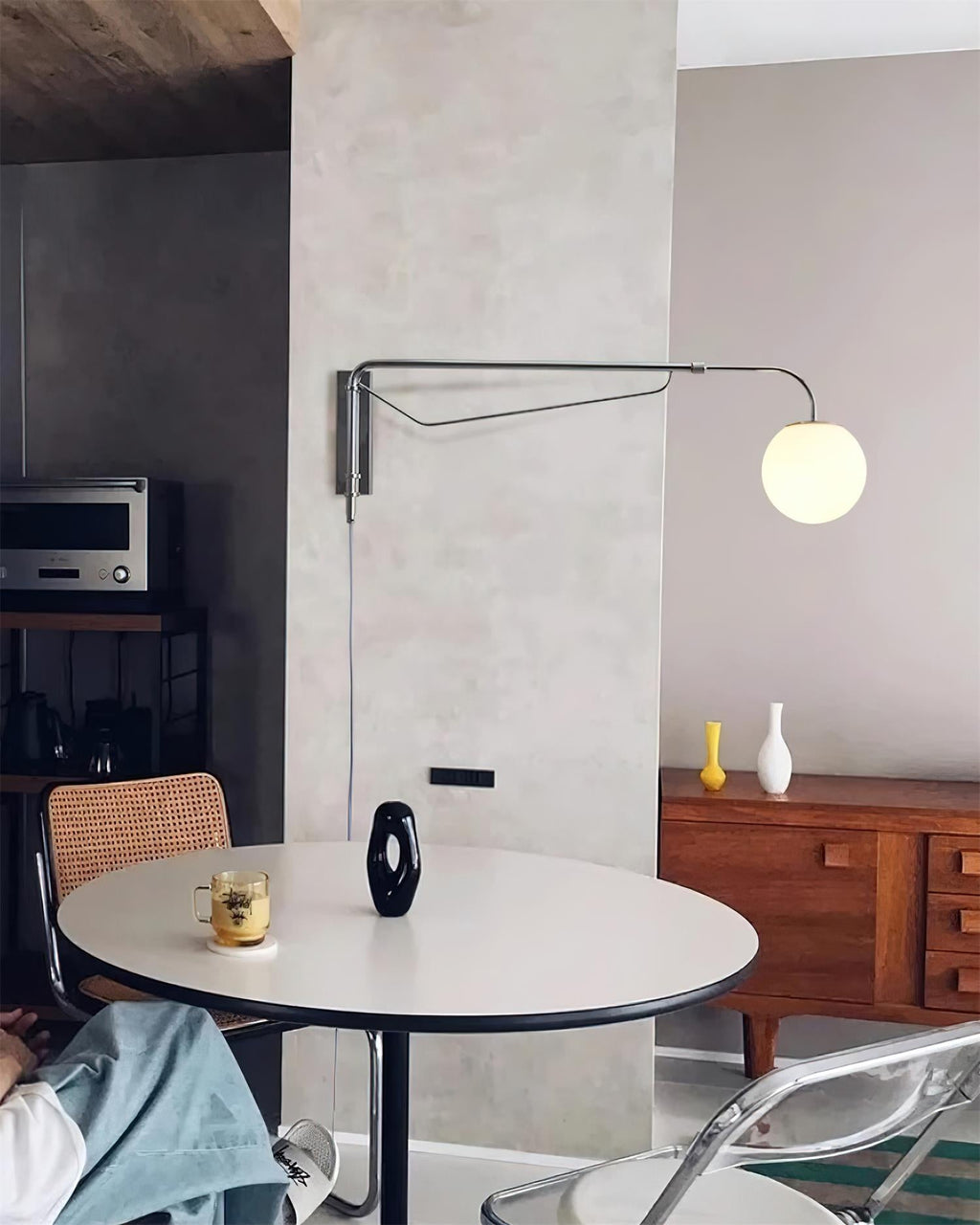 A chrome Clérachic wall light with a white globe on a pale concrete-look wall, above a round white table and a walnut sideboard.