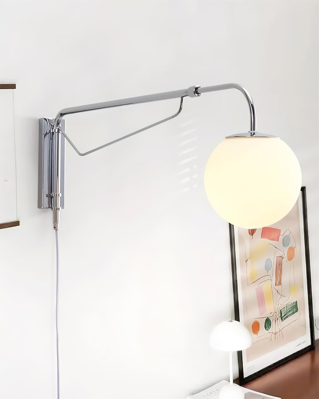 Chrome Clérachic wall light swung out to the left of its mounting plate, with a lit white globe above a framed print and a small lamp.