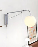 Chrome Clérachic wall light swung out to the left of its mounting plate, with a lit white globe above a framed print and a small lamp.