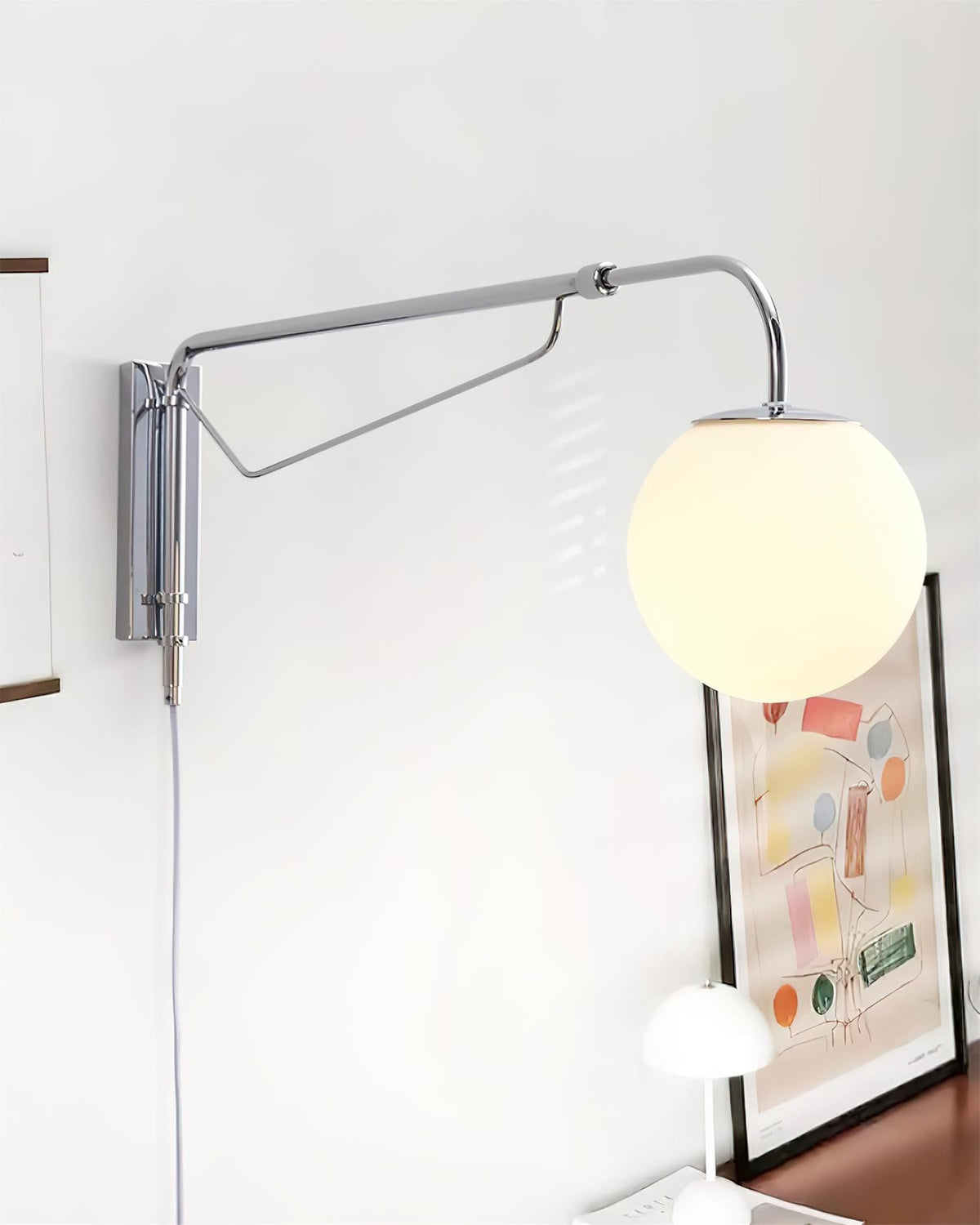 Chrome Clérachic wall light swung out to the left of its mounting plate, with a lit white globe above a framed print and a small lamp.
