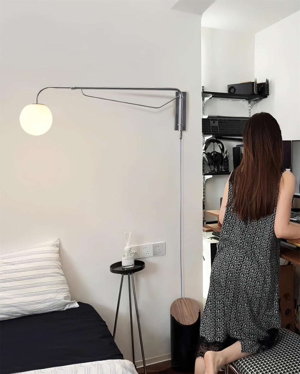 A chrome Clérachic wall light with a small white globe in a compact room, above a bed corner, with a person standing at a desk.