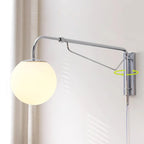 Chrome Clérachic wall light with a lit white globe by a beige curtain, shown with a green arrow marking its swivel at the wall plate.