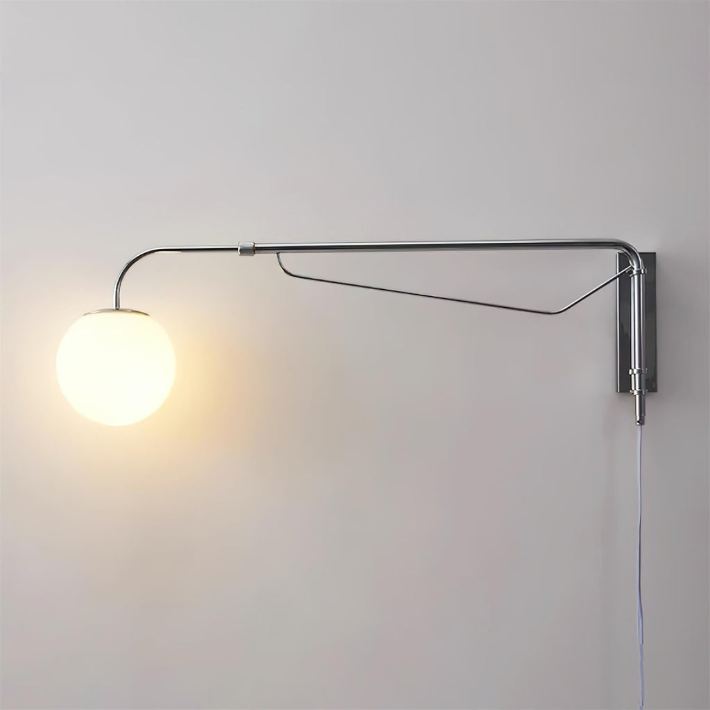 A chrome Clérachic wall light with a lit white globe glowing warm on a pale grey wall, with a narrow braced arm and a hanging cord.