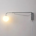 A chrome Clérachic wall light with a lit white globe glowing warm on a pale grey wall, with a narrow braced arm and a hanging cord.