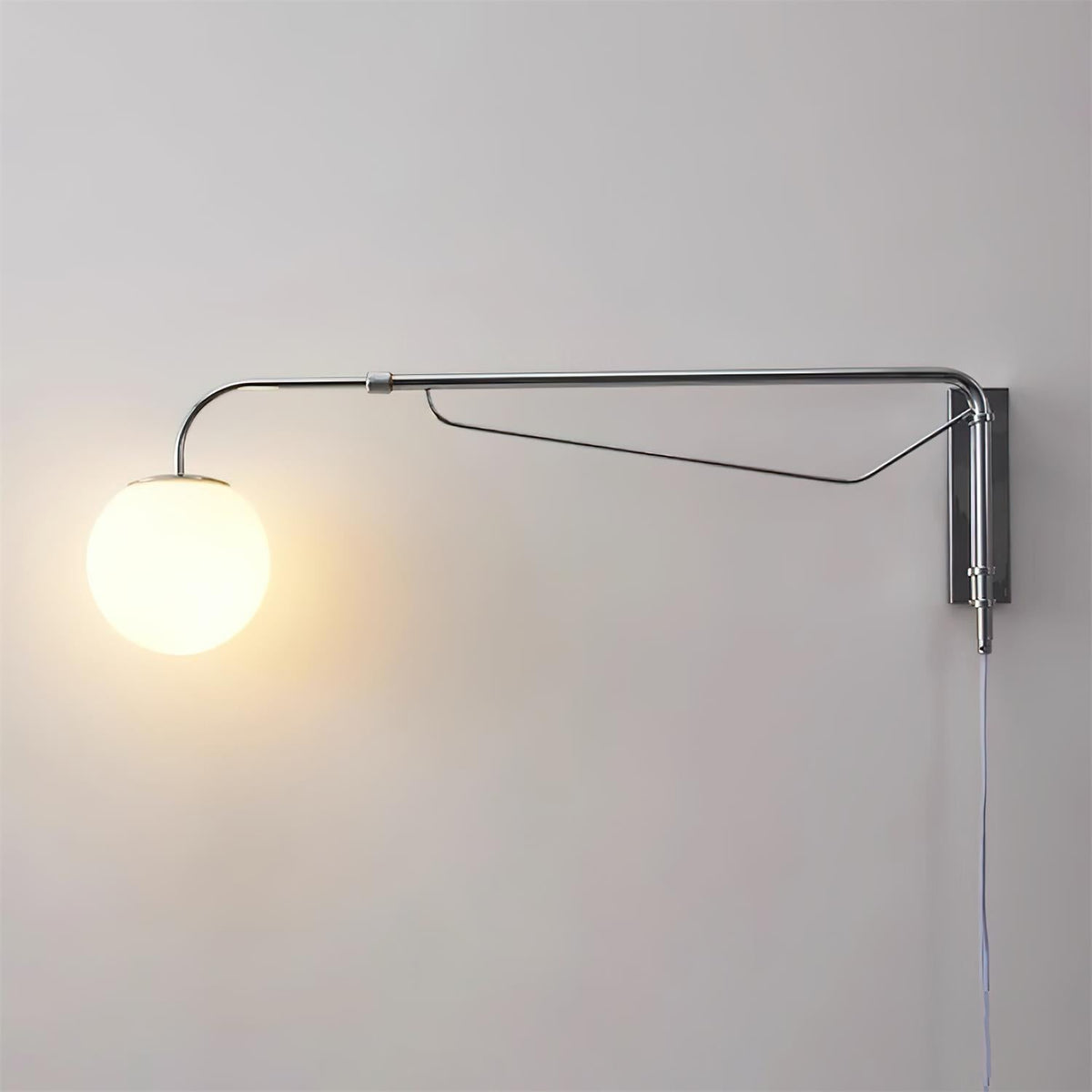 A chrome Clérachic wall light with a lit white globe glowing warm on a pale grey wall, with a narrow braced arm and a hanging cord.