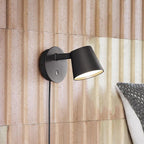 Lit black Clérachic wall light against a ribbed blush-pink panelled wall, its cord running down beside a grey cushion.