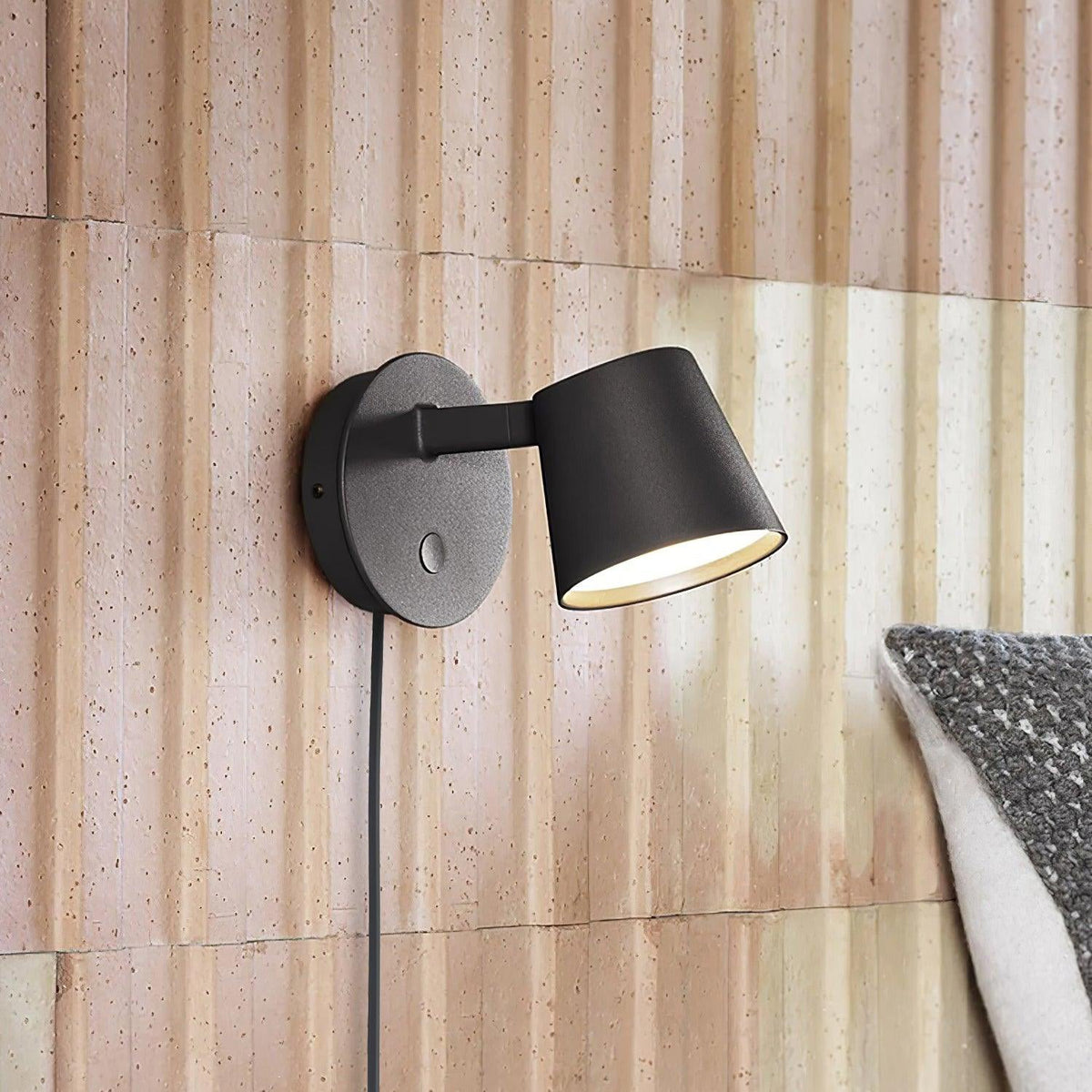 Lit black Clérachic wall light against a ribbed blush-pink panelled wall, its cord running down beside a grey cushion.