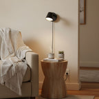 Small black Clérachic wall light above a round wooden side table, with a cream throw over a sofa arm nearby.