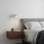 White Clérachic wall light with a slim cord above a walnut floating bedside unit, beside a grey upholstered headboard.