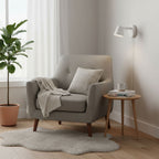 White Clérachic wall light beside a grey armchair and a potted fig, with a pale sheepskin rug on the floor.
