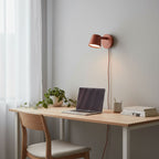 Red Clérachic wall light above a light wood desk with a laptop, a trailing plant and a stack of books.