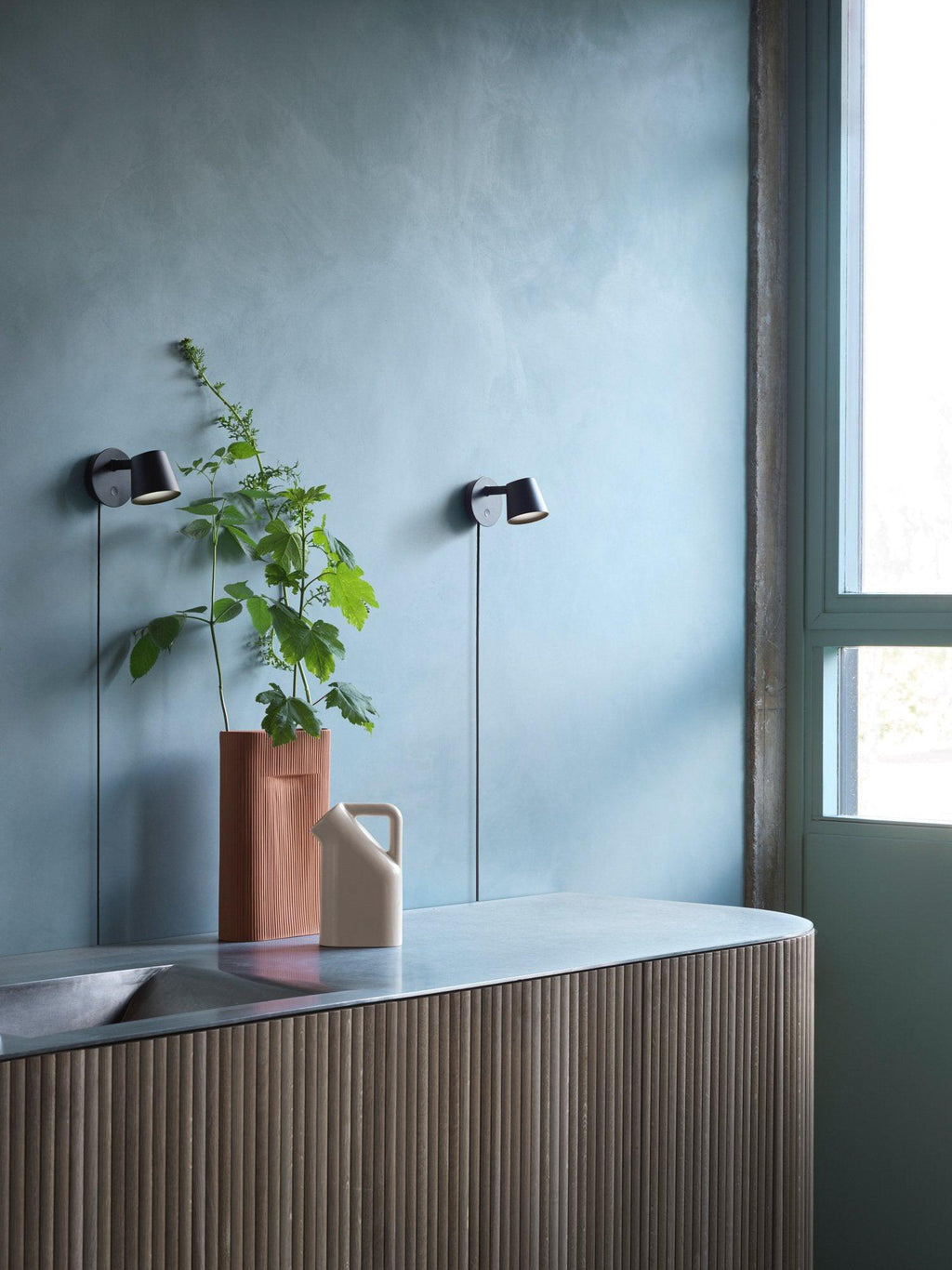 Clérachic black wall lights seen across a room with a blue plaster wall, a leafy plant and a ribbed sideboard.