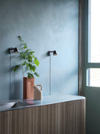 Clérachic black wall lights seen across a room with a blue plaster wall, a leafy plant and a ribbed sideboard.