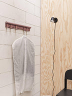 Black Clérachic wall light and a clothes hook rail on a pale brick wall beside a plywood panel, with a garment bag hanging.
