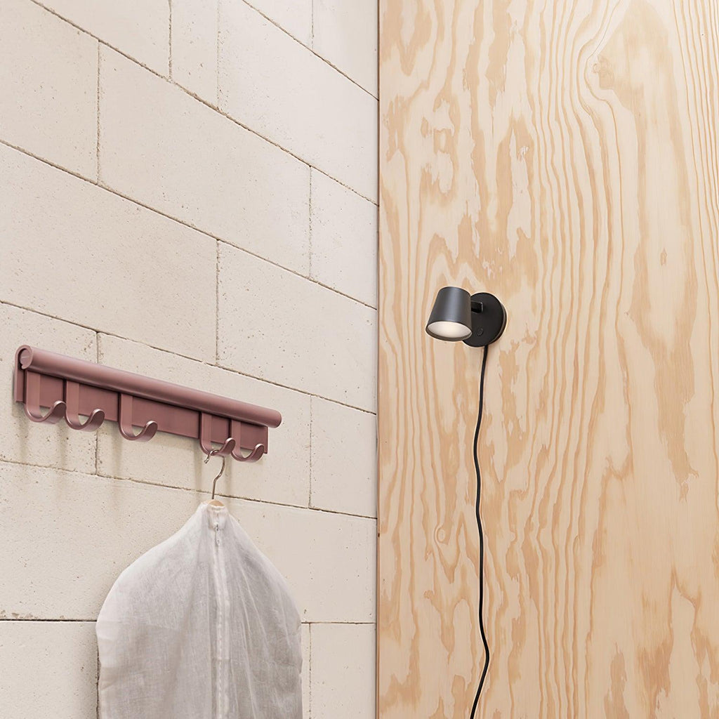 Black Clérachic wall light with its cord trailing down a plywood panel, next to a pink hook rail holding a garment bag.