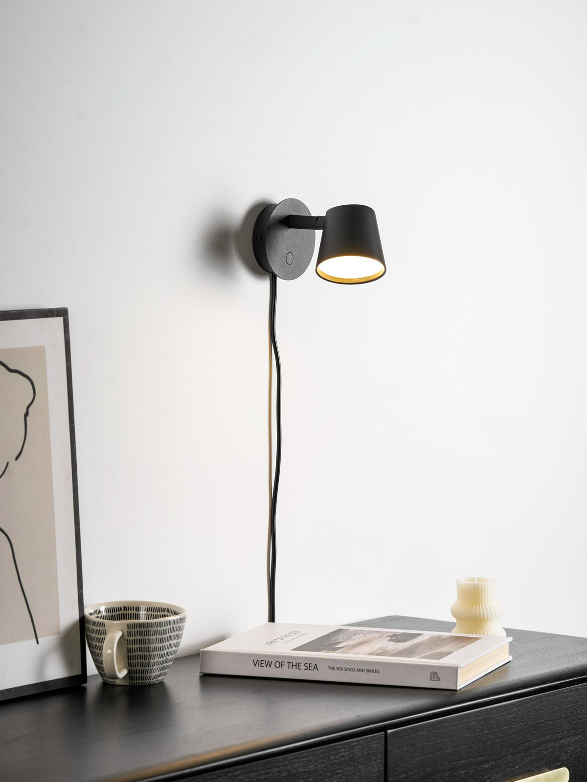 Black Clérachic wall light on a white wall above a dark chest of drawers, with a book, a mug and a ribbed candle.