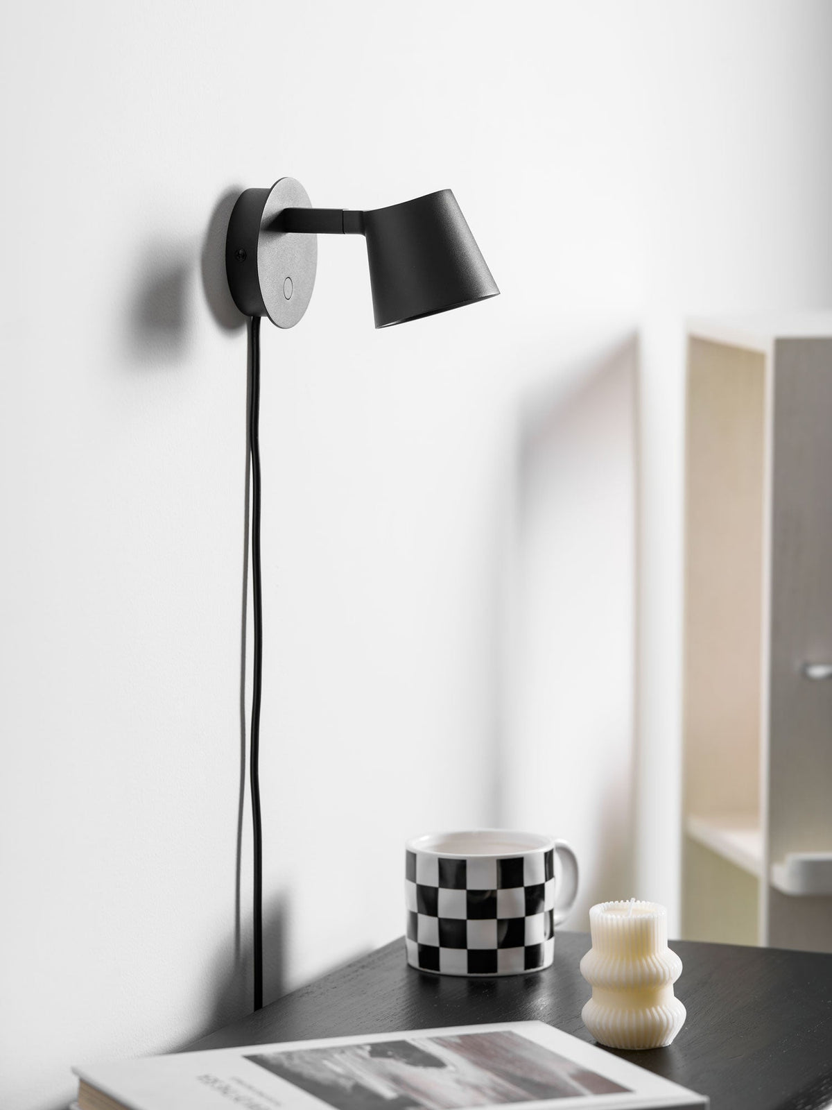 Side-on view of a black Clérachic wall light with a long cord, above a checkered mug and a ribbed candle on a dark cabinet.