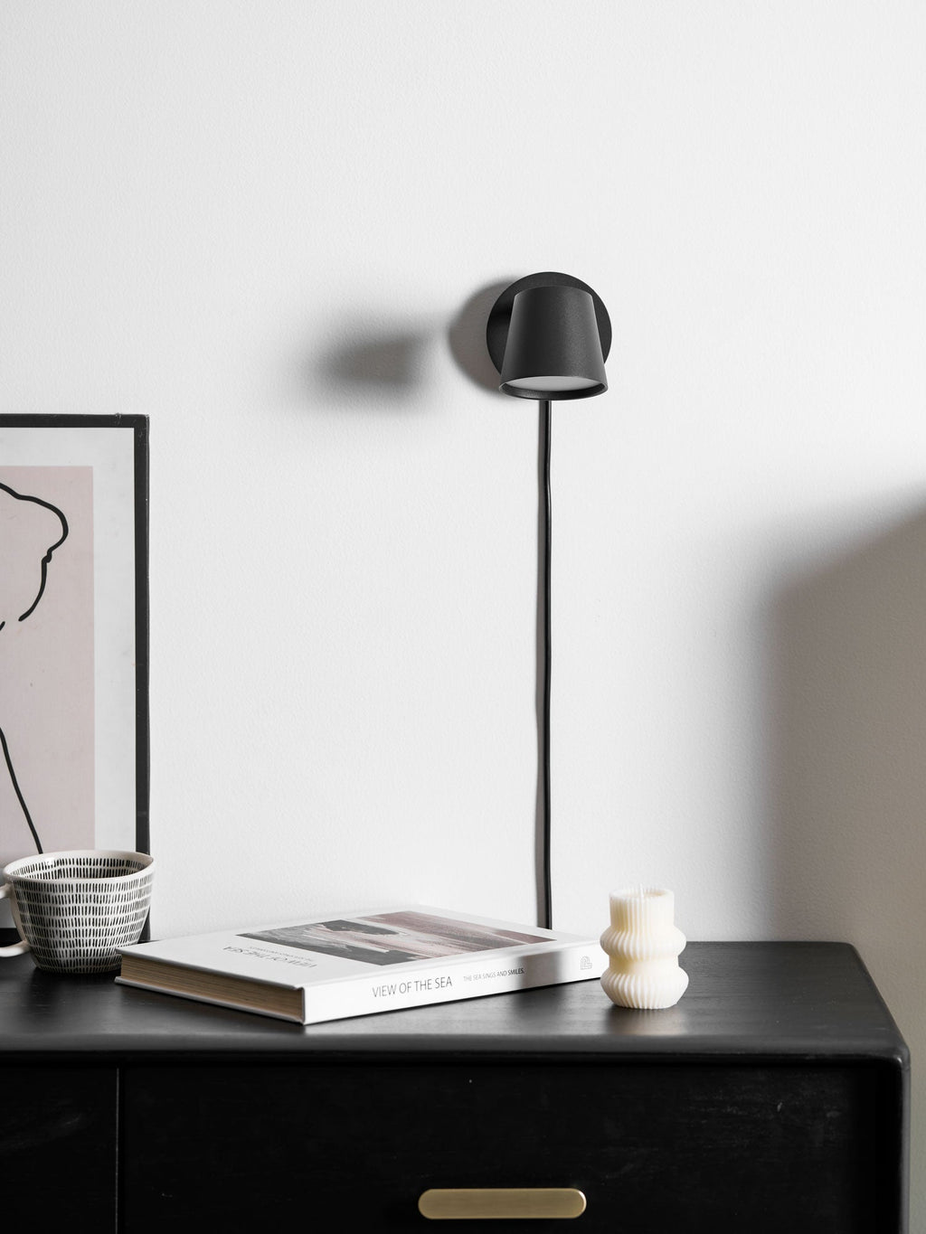 Black Clérachic wall light with its shade turned down, cord running to a dark cabinet topped with a book and a striped mug.