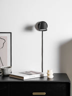Black Clérachic wall light with its shade turned down, cord running to a dark cabinet topped with a book and a striped mug.