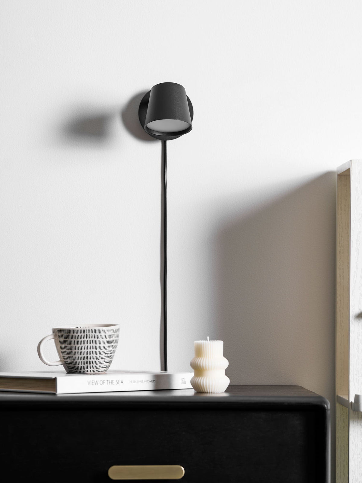 Black Clérachic wall light angled to face the wall, with a book, a ribbed candle and a mug on the cabinet beneath.