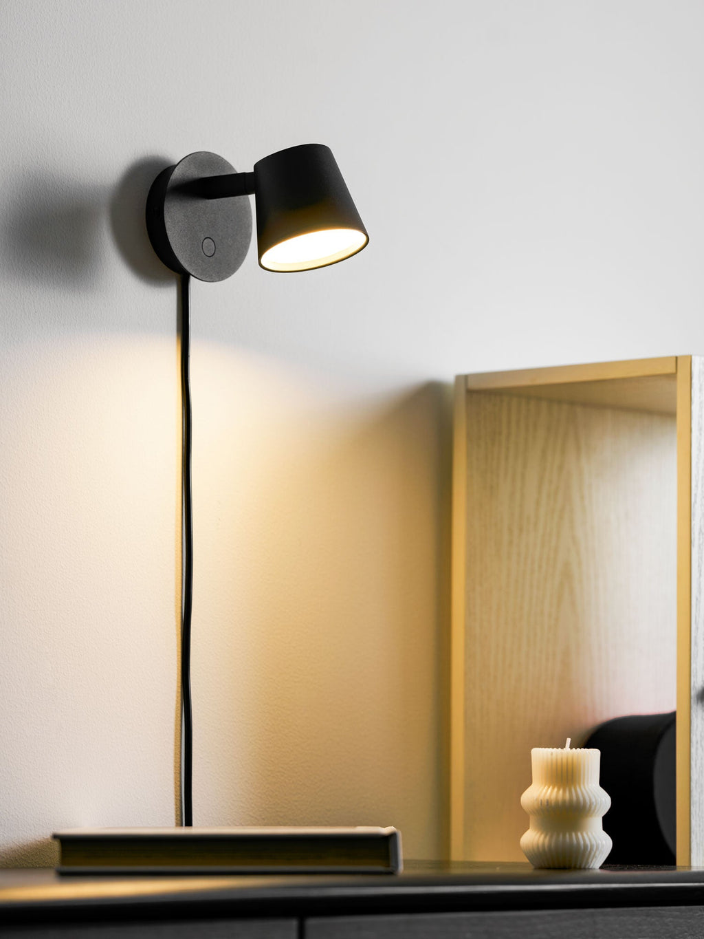 Lit black Clérachic wall light casting a warm pool of light down a white wall, with a pale wooden frame behind.