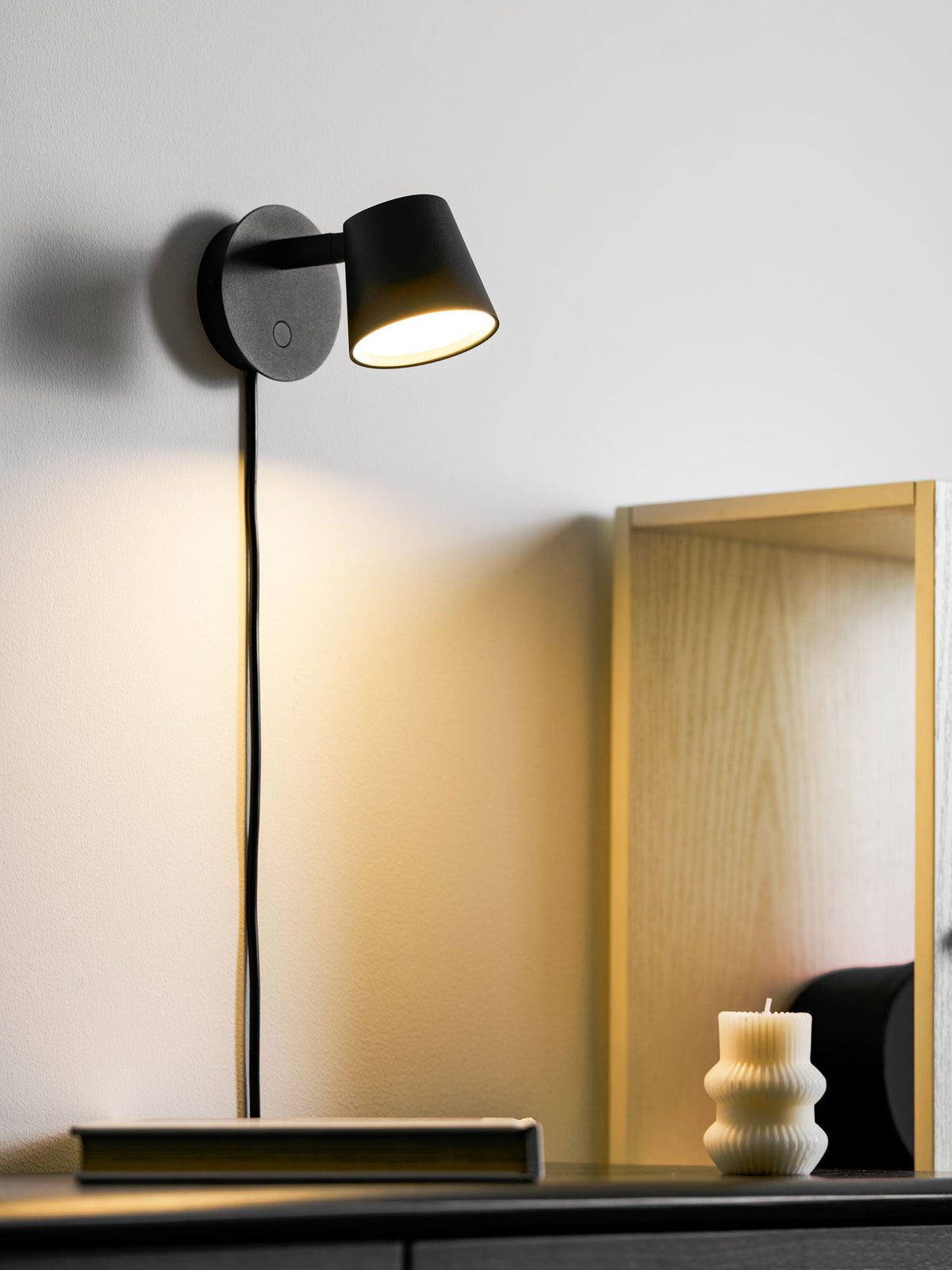 Lit black Clérachic wall light casting a warm pool of light down a white wall, with a pale wooden frame behind.