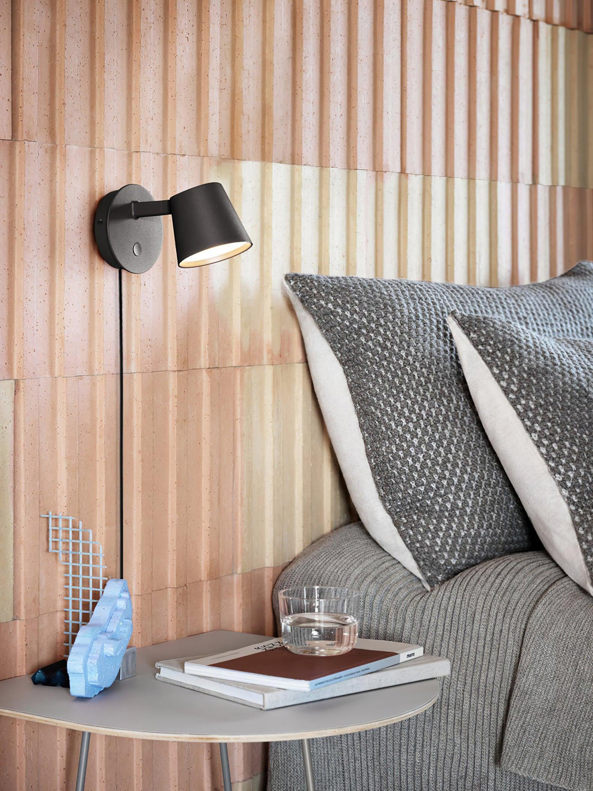 Black Clérachic wall light above a bedside table, warm light spilling across blush ribbed panelling and grey bedding.