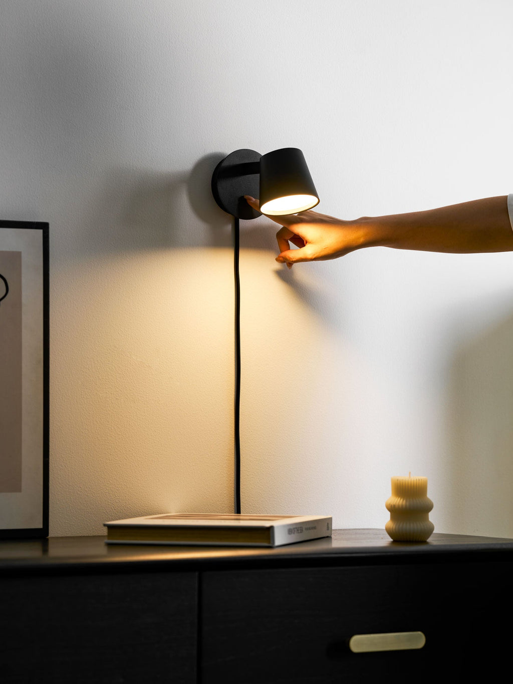 Hand reaching to press the switch on a lit black Clérachic wall light, warm light falling over the wall below.