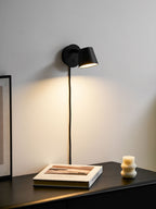 Black Clérachic wall light switched on above a dark cabinet, giving a soft cone of warm light on the white wall.