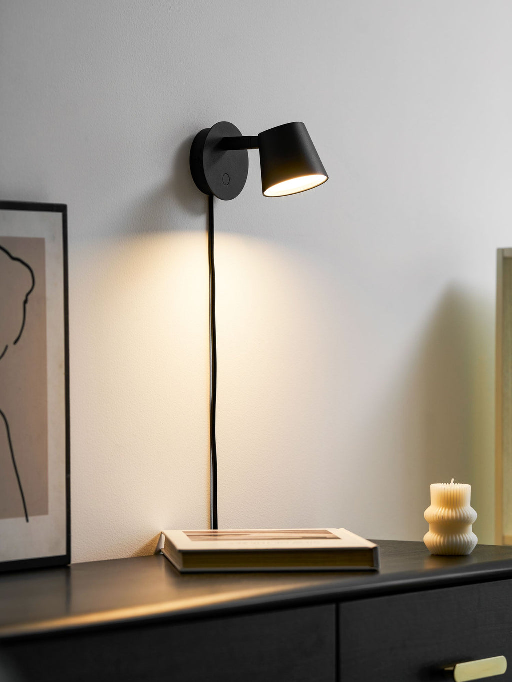 Lit black Clérachic wall light beside a framed print, a book and a candle on a dark chest of drawers.