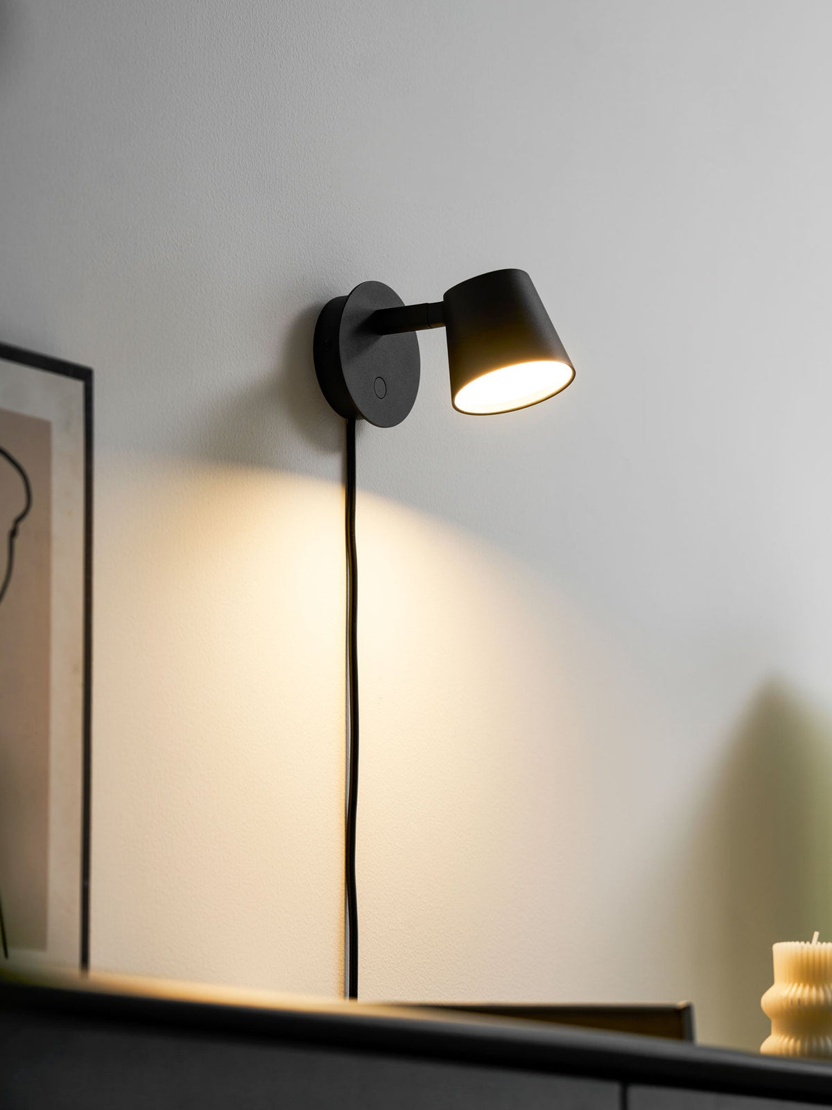 Lit black Clérachic wall light on a white wall, its warm glow falling over a framed print and the edge of a dark cabinet.