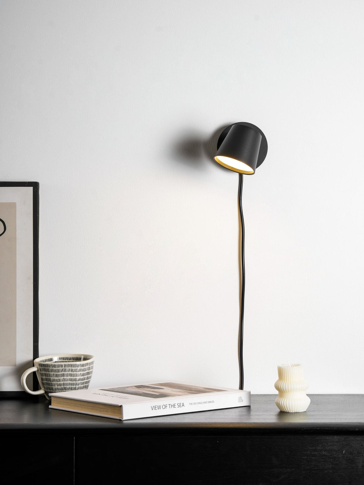 Black Clérachic wall light with its shade turned down, a cord hanging to a book and a striped mug on a dark cabinet.